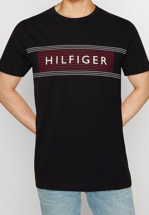 Osoba w czarnej koszulce z czerwonymi i białymi poziomymi paskami oraz napisem "HILFIGER" na piersi, w połączeniu z jasnoniebieskimi jeansami.
