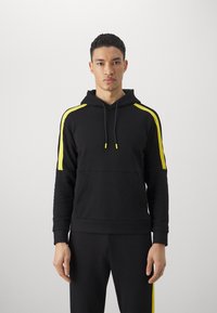 Felpa nera con strisce gialle sulle maniche, cappuccio con cordino e polsini a coste. Tessuto liscio con un design aderente. Pantaloni neri coordinati.