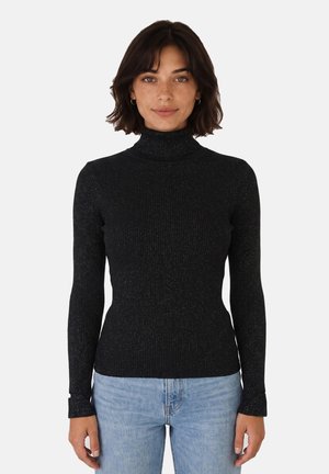Donna con capelli corti e scuri che indossa un maglione nero a collo alto a maniche lunghe e jeans azzurri, in piedi davanti a uno sfondo bianco semplice.