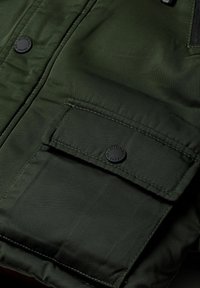 Veste en tissu vert à motif avec des côtes verticales, dotée d'une grande poche à rabat et d'une fermeture à bouton-pression noir. Détails de surface texturée visibles.