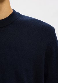 Krupan kadar osobe koja nosi tamnoplav, pleteni džemper s okruglim izrezom na jednobojnoj svijetloj pozadini.