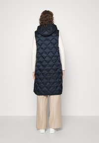 Persona vestida con un chaleco acolchado negro sin mangas y con capucha, pantalones beige de pierna ancha y zapatos blancos, de pie frente a un fondo liso.