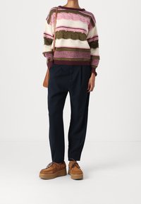 Person i en randig stickad tröja med rosa, krämfärgade och olivfärger, marinblå tapered-byxor, bruna platå snörskor och en brun axelväska.