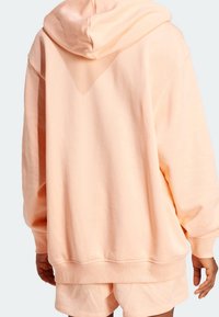Persikofärgad hoodie med en rymlig passform, justerbar huva och ribbade ärmslut, tillverkad av mjukt tyg. Synlig sömnad och detaljer vid fållen.