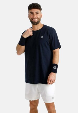 Lächelnder Mann mit schwarzem T-Shirt, weißen Shorts und schwarzen Handgelenksbändern, der vor einem schlichten hellen Hintergrund steht.