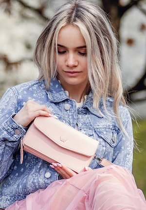 Pink faux leather wallet with a flap closure, featuring a logo detail. The wallet is held against a denim jacket with a paisley pattern.