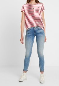 Camiseta a rayas rojas y blancas de manga corta con un pequeño detalle gráfico, combinada con jeans ajustados de color azul claro y zapatillas blancas.