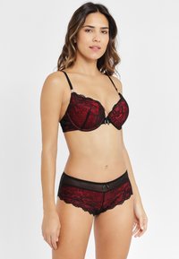 Roter und schwarzer Spitzen-BH mit Unterdrahtunterstützung, der detaillierte Blumenmuster aufweist, verstellbare Träger und eine dekorative Schleife in der Mitte vorne hat. Passende Spitzen-Boyshorts mit einem durchsichtigen Einschnitt und gewellten Kanten.