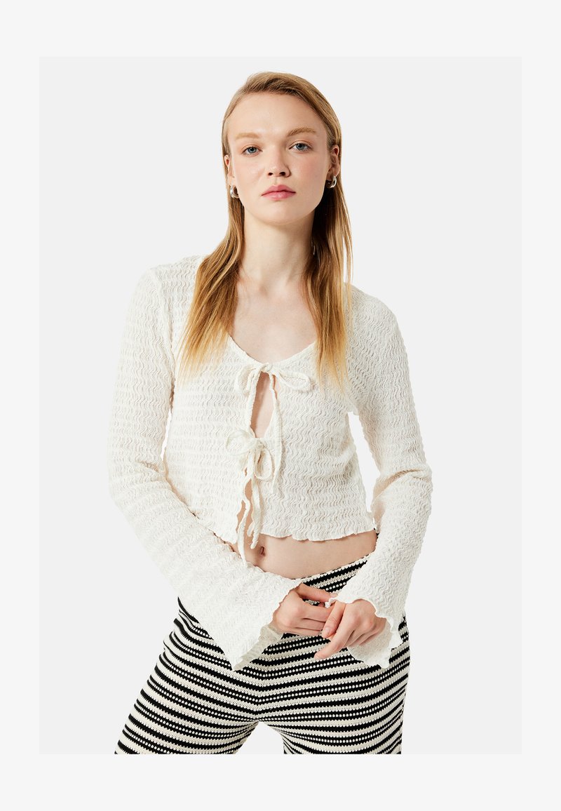 Joven mujer con cabello rubio largo que lleva un cárdigan blanco texturizado corto con lazos y pantalones a rayas en blanco y negro, de pie contra un fondo blanco.