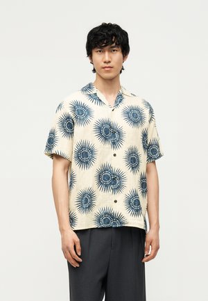 Giovane uomo che indossa una camicia beige a maniche corte con bottoni e motivi a raggiera blu e pantaloni grigio scuro, in piedi davanti a uno sfondo semplice.