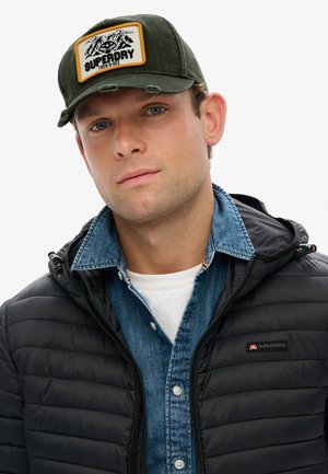 Groene pet met een patch met bergen en tekst, gedragen met een zwarte pufferjas en een spijkerhemd, wat een casual gelaagde stijl laat zien.