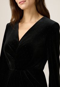 Abito in velluto nero con profondo scollo a V, maniche lunghe e un corpetto aderente, decorato con piccoli dettagli luccicanti.