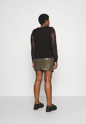 Femme debout, tournée de dos, portant un haut noir avec des manches en dentelle florale transparente et une mini-jupe en similicuir vert olive avec des chaussures noires.
