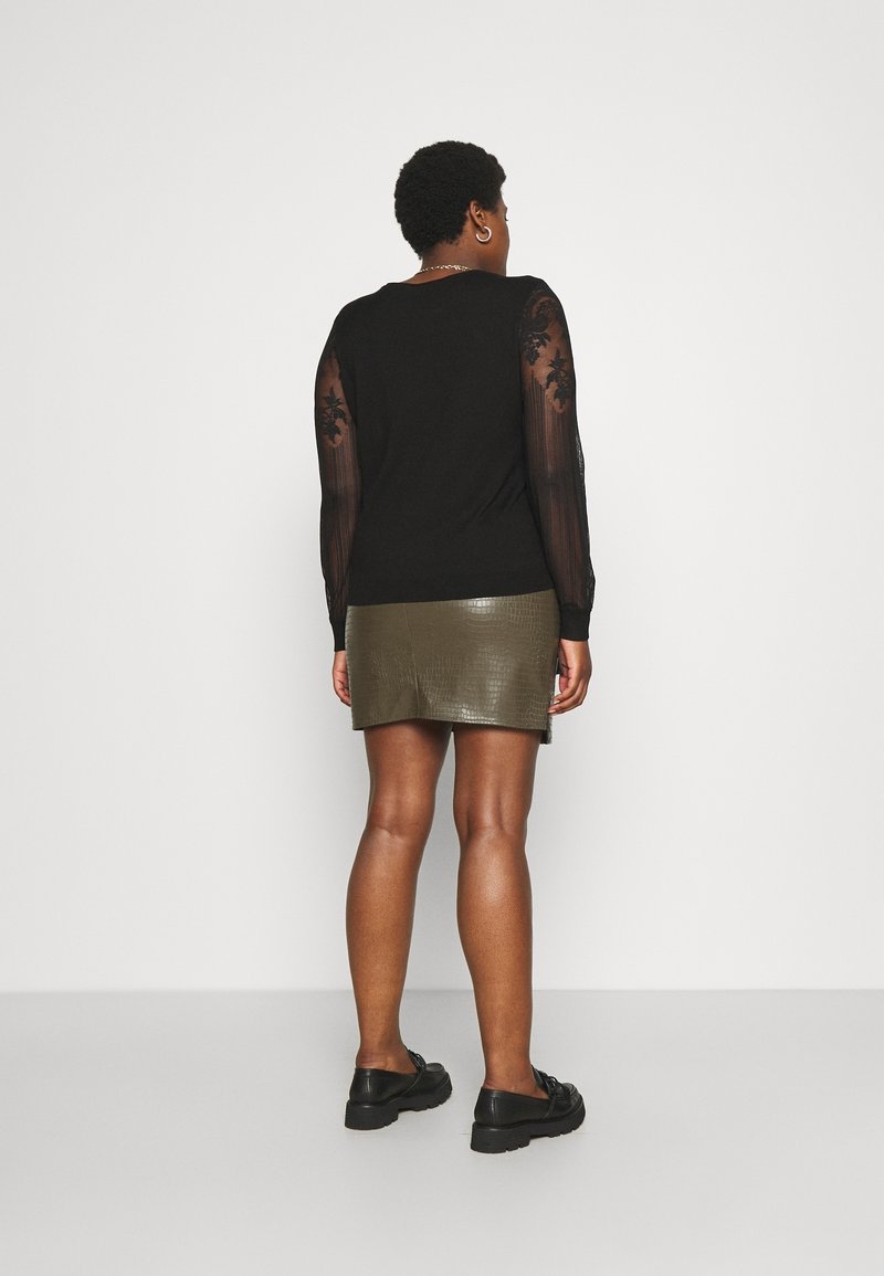 Femme debout, tournée de dos, portant un haut noir avec des manches en dentelle florale transparente et une mini-jupe en similicuir vert olive avec des chaussures noires.
