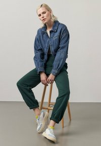 Giacca in denim, pantaloni verdi e scarpe da ginnastica grigie con accenti gialli. Modello seduto su uno sgabello di legno contro uno sfondo chiaro.