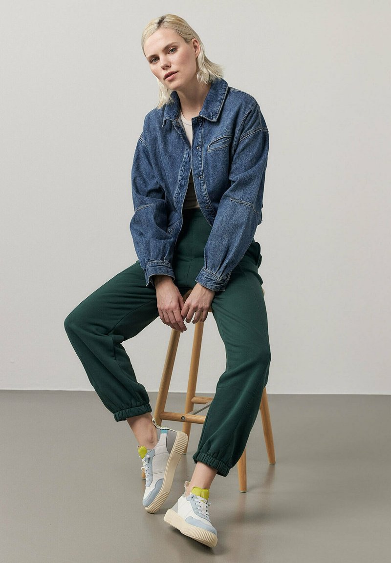 Jeansjacke, grüne Hose und graue Sneakers mit gelben Akzenten. Modell sitzt auf einem Holzstuhl vor einem hellen Hintergrund.