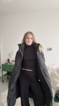 Μαύρο φουσκωτό παλτό με ψηλό γιακά, φορεμένο πάνω από ένα μαύρο κοντό μπλουζάκι και μαύρο παντελόνι. Το υλικό φαίνεται γυαλιστερό και διαθέτει μεγάλα μπροστινά τσέπες.