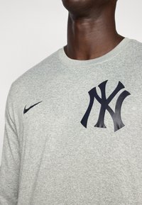 Lys grå langermet t-skjorte med rund hals, med et stort svart New York Yankees-logo på brystet og et lite svart Nike-logo på skulderen.