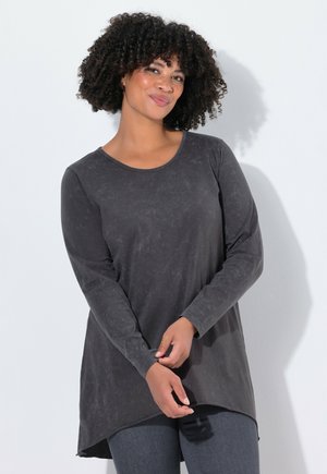 Langärmlige, graue Tunika mit Rundhalsausschnitt und asymmetrischem Saum. Glatte Textur mit einem subtilen marmorierten Muster. Kombiniert mit dunklen Jeans.