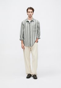 Hombre joven de pie, vistiendo una camisa de botones a rayas grises, pantalones de pierna ancha color crema y mocasines marrón oscuro, frente a un fondo blanco liso.