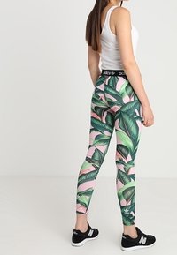 Legíny s růžovým pozadím s zelenými tropickými listovými vzory, s vysokým pasem a černým logem, vyrobené z elastického materiálu.