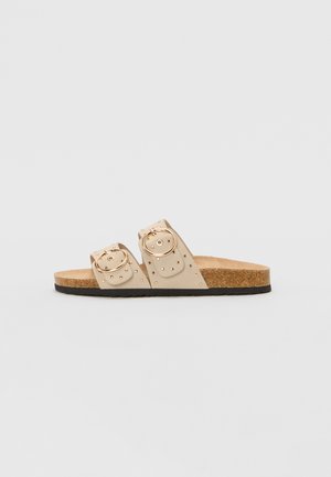 Beige slide sandal with two wide straps featuring circular metallic buckles and studded accents, set on a cork footbed with a black outsole.