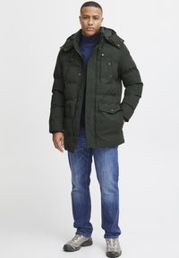 Chaqueta puffer de color verde oscuro con capucha, cierre de cremallera, múltiples bolsillos y textura suave. Llevada sobre un jersey de cuello alto azul marino y vaqueros azules.