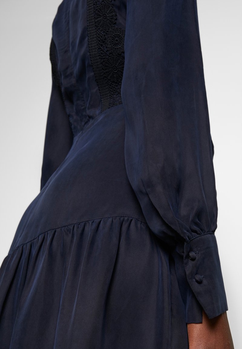Mörkblå klänning med långa ärmar, med spetsdetaljer på bodicen och knappdekorationer vid ärmsluten, mjukt och flödande tyg, plisserad kjol.