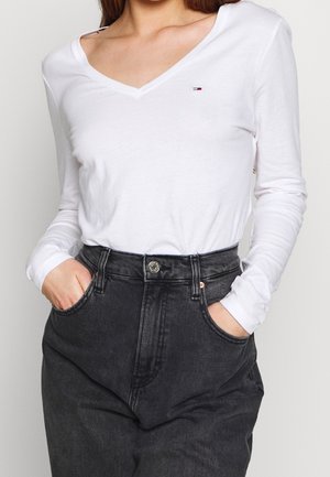 T-shirt bianca a maniche lunghe in cotone con scollo a V e piccolo logo ricamato, abbinata a jeans in denim grigio scuro a vita alta con tasche anteriori.