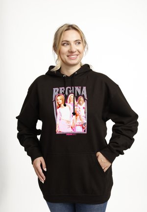 Žena se usmívá, má na sobě černou mikinu s růžovým nápisem „Regina“ a „Mean Girls“ a grafikou čtyř ženských postav na přední straně.