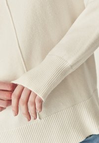 Crèmewitte trui met een zachte textuur, geribbelde manchetten en een losse pasvorm. Zichtbare stikdetails langs de zoom.