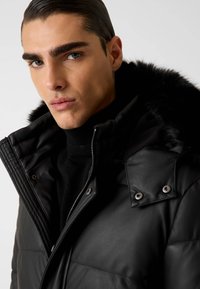 Giacca nera imbottita con cappuccio in ecopelliccia, caratterizzata da chiusura con zip e bottoni a pressione, dettagli a coste sul colletto e accenti in metallo.