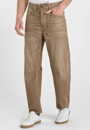 Brown loose-fit pants with large front pockets and drawstring waist worn with white sneakers on a standing person.