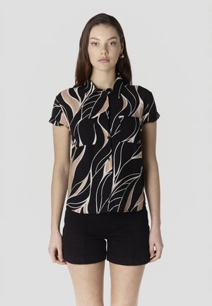 Noor naine kannab lühikeste varrukatega nööbitavat särki, millel on musta ja beeži abstraktse lehemustriga kujundus, ning musti lühikesi pükse, seisab ühevärvilise tausta ees.