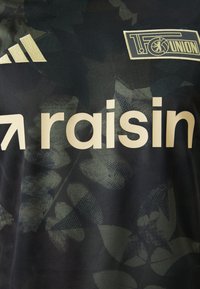 Tumši zaļš futbola krekls ar lapu rakstiem, ar baltu logotipu un "raisin" tekstu. Izgatavots no elpojoša auduma, iekļauj Adidas svītras.