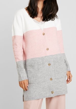 Kvinde iført en lang cardigan med knapper, i hvid, pink og grå farveblokke, med en blonde detalje på brystet og forlommer.