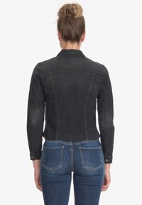 Kvinna med blont hår i en knut, iklädd en figursydd svart denimjacka och blå jeans, vänd bort från kameran mot en enkel vit bakgrund.