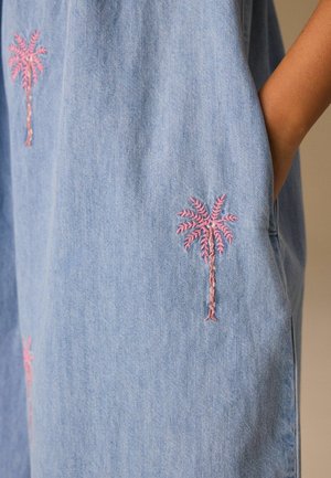 Hellblauer Denimstoff mit rosa bestickten Palmen, auf dem der Arm einer Person zu sehen ist, deren Hand in einer Seitentasche ruht.