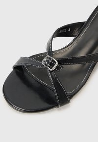 Sandale en cuir noir avec un design entrecroisé, une texture brillante, des accents de couture blanche et un détail de boucle en métal sur la sangle.
