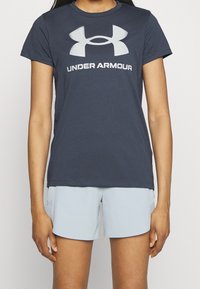Mörk marinblå t-shirt med kort ärm och en stor ljusgrå "Under Armour"-logotyp på framsidan. Matchas med ljusblå shorts. Slät textur.