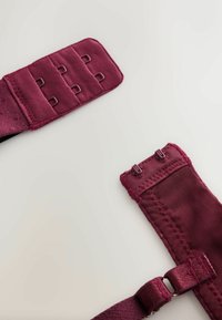 Close-up van een bordeauxrood bh-bandje met haak-en-oogsluiting op een witte achtergrond, waarbij de haken en verstelbare stoffen delen zichtbaar zijn.