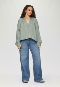 Hellgrüne Bluse mit langen Ärmeln und V-Ausschnitt, gerafften Schultern und elastischen Bündchen, kombiniert mit weit geschnittenen blauen Jeans.