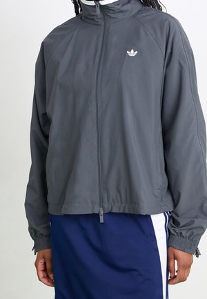 Personne portant une veste zippée gris foncé avec un logo Adidas blanc et une jupe bleue avec des bandes blanches et bleues sur les côtés.