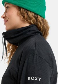 Čierna fleecová bunda s vysokým golierom, so zipsom na krku, pružnou textúrou a bielym logom "ROXY" na rukáve. Na hlave zelená čiapka.