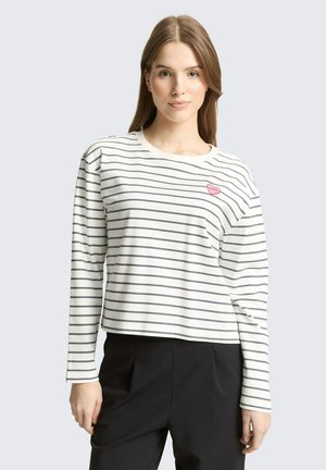 Jeune femme portant une chemise blanche à manches longues avec de fines rayures horizontales noires et un petit détail en forme de cœur rose, associée à un pantalon noir.