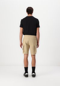 Beige Cargo-Shorts mit Seitentaschen, elastischem Bund und lockerer Passform, kombiniert mit einem schwarzen T-Shirt mit kurzen Ärmeln und schwarzen Socken.