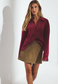 Vrouw met lang blond haar, gekleed in een bordeauxrode blouse met kantdetails en een olijfgroene rok, staande voor een witte achtergrond met schaduw.
