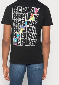 Camiseta negra con el texto "REPLAY" en blanco contorneado repetido, algunas letras llenas de patrones abstractos coloridos, llevada por una persona con jeans azules.