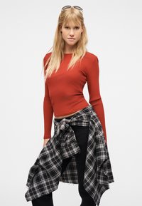 Jeune femme aux longs cheveux blonds, portant un haut rouge à manches longues, un pantalon noir, une chemise à carreaux nouée autour de la taille, et des lunettes de soleil sur la tête.