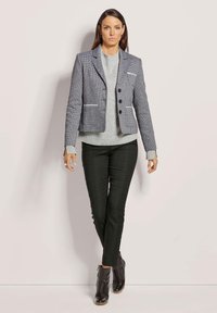 Maßgeschneiderte Blazer im Hahnentrittmuster, grauer Stoff, einreihig mit drei Knöpfen, kombiniert mit einem hellgrauen Pullover und schlanken schwarzen Hosen.